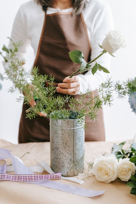 A florist wearing a white top and brown apron arranges a floral bouquet in a tall, cylindrical, silver-grey metallic vase. The bouquet features a single large white rose held delicately by the florist