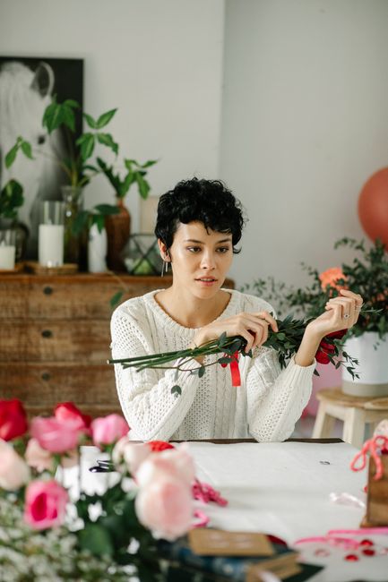 A woman with short, curly black hair and light skin is arranging a bouquet of fresh flowers at a table. She is wearing a white knitted sweater and is focused on her task with a thoughtful expression. 