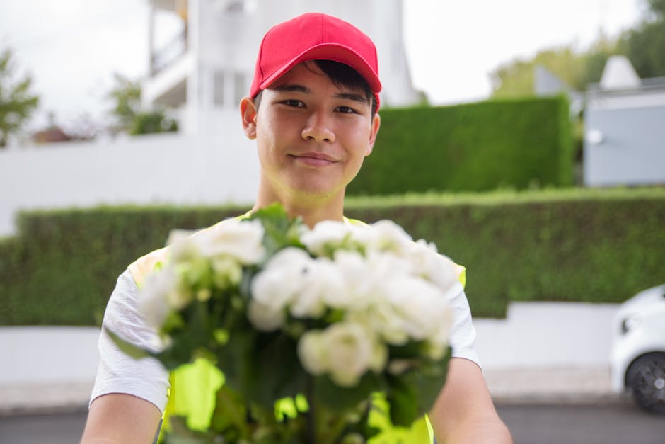 A smiling delivery person wearing a red cap and a neon yellow safety vest is holding a bouquet of fresh white flowers, possibly roses or peonies, with green foliage. The bouquet is wrapped in clear pl