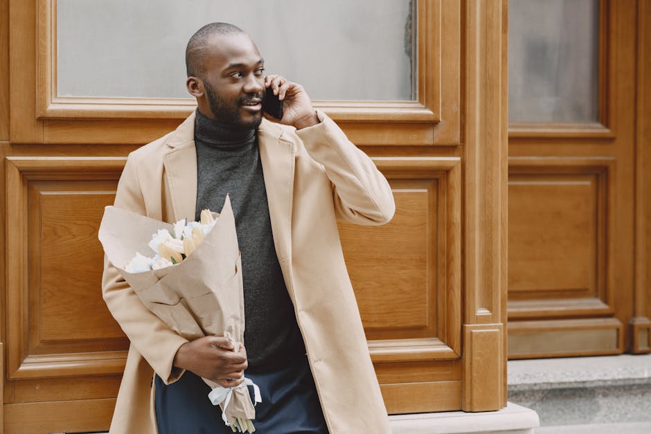 A man with dark skin, a neatly groomed beard, and a shaved head, dressed in a beige overcoat over a dark grey turtleneck sweater, is standing outdoors near a wooden-paneled building. He is holding a b